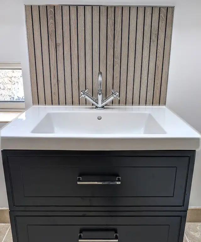 Dark vanity unit with white basin in front of a wood slat effect splashback